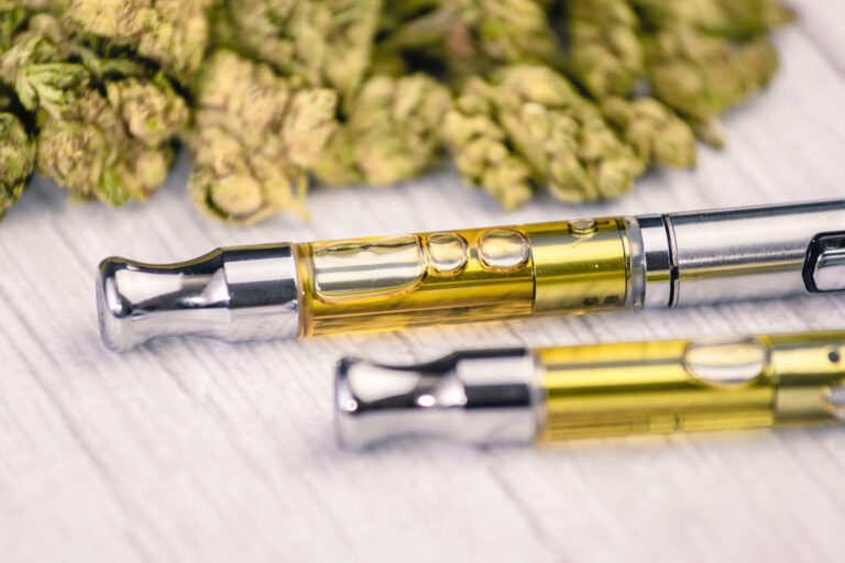 THC/CBD cannabis vape pens up-close with marijuana leaf on white wooden table